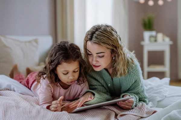 Gelukkig moeder met haar dochtertje liggend op bed en met behulp van tablet thuis. — Stockfoto