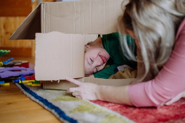 เด็กชายที่มีอาการดาวน์กับแม่ของเขาเล่นกับกล่องด้วยกันที่บ้าน . — ภาพถ่ายสต็อก