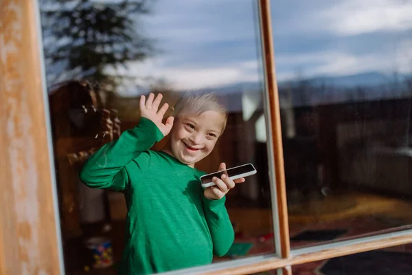Glücklicher kleiner Junge mit Down-Syndrom nutzt Smartphone und winkt durch Fenster — Stockfoto