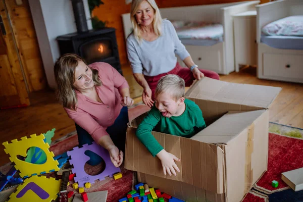 Niño con síndrome de Down con su madre y su abuela jugando con la caja juntos en casa. — Foto de Stock