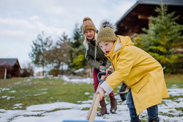 Pojke med Down syndrom med sin mor som rensar snö från vägen med spade framför huset. — Stockfoto