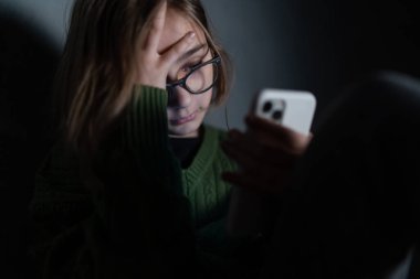 Üzgün küçük kız, karanlıkta yalnız, oturuyor ve akıllı telefon kullanıyor..