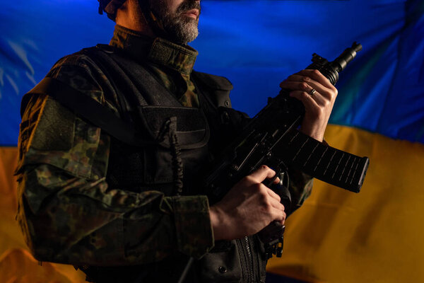Close-up of soldier in military uniform holding weapon with Ukraine flag in background.