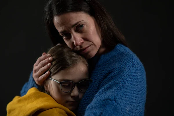 Smutná matka objímající svou dceru, oba na sobě ukrajinské národní barvy na černém pozadí. — Stock fotografie