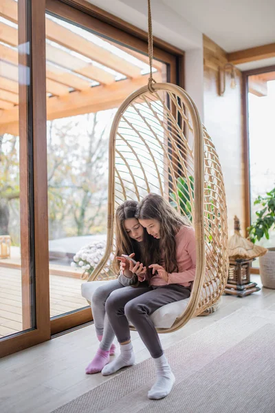 Glada små systrar sitter i rotting rottingstol och använder smartphone inomhus i uterum. — Stockfoto