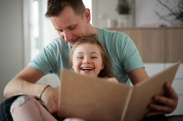 Padre che legge un libro alla sua figlioletta con la sindrome di Down quando è seduto sul letto a casa. — Foto Stock