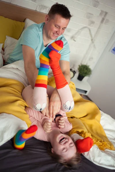Niña alegre con síndrome de Down acostada en la cama y riendo cuando su padre le hace cosquillas. — Foto de Stock