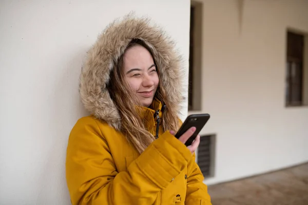 ダウン症候群を持つ幸せな若い女性がパーカーを身に着けています,壁を傾け、スマートフォンを使用して. — ストック写真