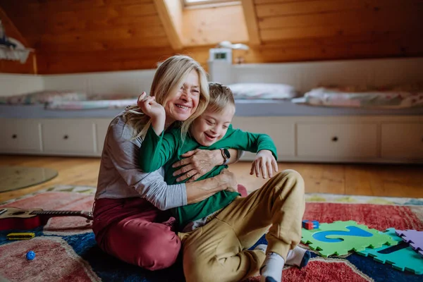 Happy boy met Down syndroom zitten op de vloer en knuffelen met zijn grootmoeder thuis. — Stockfoto
