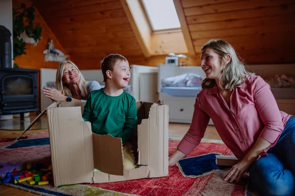 Ragazzo con la sindrome di Down con la madre e la nonna che giocano con la scatola insieme a casa. — Foto Stock