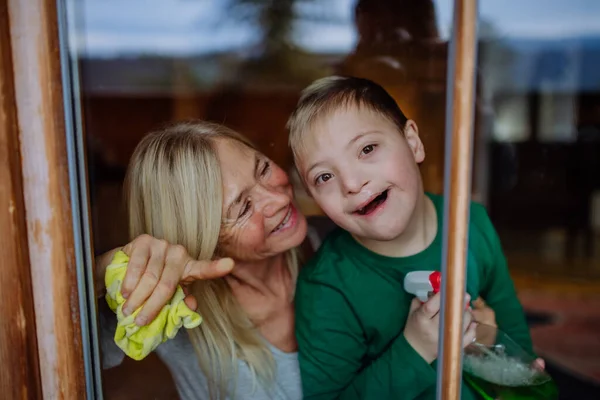 Junge mit Down-Syndrom bei Großmutter beim Fensterputzen zu Hause. — Stockfoto