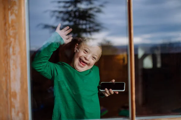 Menino feliz com síndrome de Down usando smartphone e acenando pela janela — Fotografia de Stock