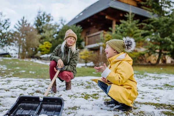 Pojke med Down syndrom med sin mor som rensar snö från vägen med spade framför huset. — Stockfoto