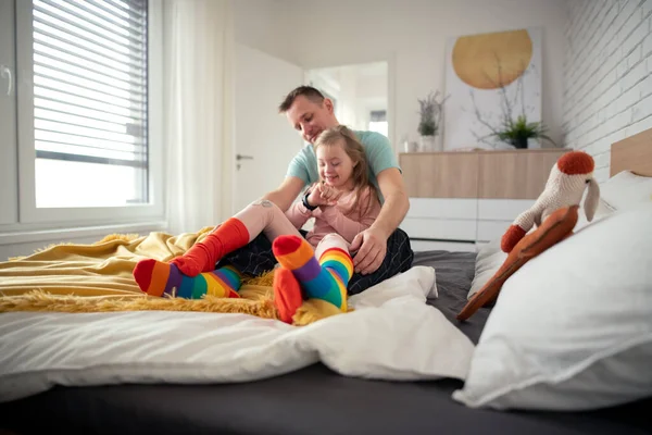 Padre che indossa calzini diversi per la sua figlioletta con la sindrome di Down quando è seduto sul letto a casa. — Foto Stock