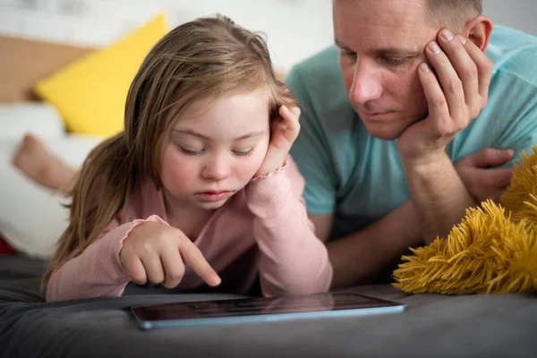 父と彼の小さな娘とともにダウン症候群ベッドの上に横たわって、自宅でタブレットを使用して. — ストック写真