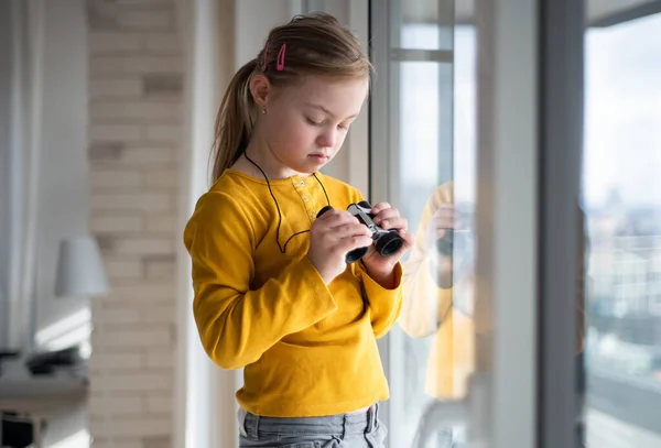 Niña curiosa con síndrome de Down con prismáticos mirando a través de la ventana en casa. — Foto de Stock