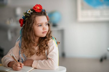 Little sad Ukrainian girl sitting in classroom during class, concept of enrolling Ukrainian kids to schools. clipart