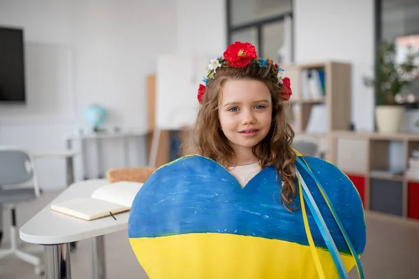 Kis ukrán iskolás lány gazdaság szív ukrán színek és nézi kamera az iskolában. — Stock Fotó