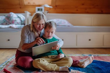Down sendromlu bir çocuk yerde oturuyor ve büyükannesiyle tablet kullanıyor..