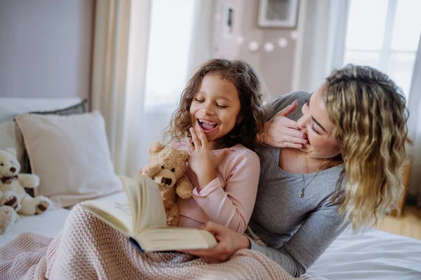 Счастливая мать со своей маленькой дочерью сидит на кровати и читает книгу дома. — стоковое фото