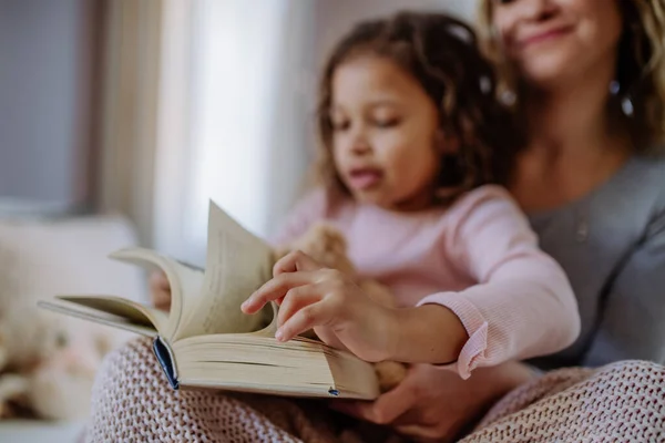 Mãe feliz com sua filhinha sentada na cama e lendo livro em casa. — Fotografia de Stock