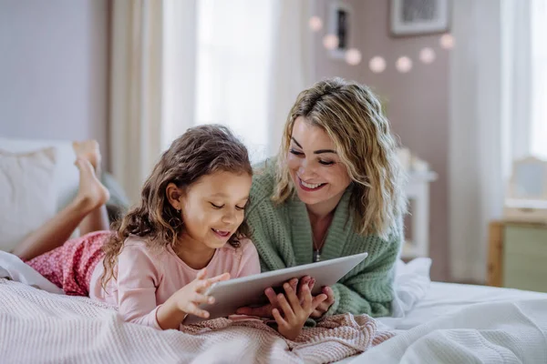 Gelukkig moeder met haar dochtertje liggend op bed en met behulp van tablet thuis. — Stockfoto