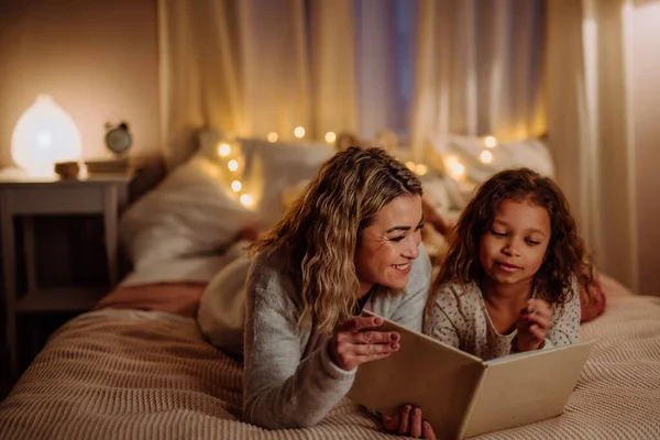 Küçük kızı yatakta yatarken ve akşam evde kitap okurken mutlu bir anne.. — Stok fotoğraf