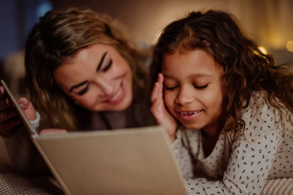 Happy mother with her little daughter lying on bed and reading book in evening at home. — Stock Photo, Image