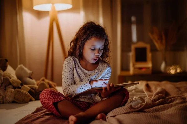Happy little multiracial girl using tablet on bed in evening at home. — Stock Photo, Image