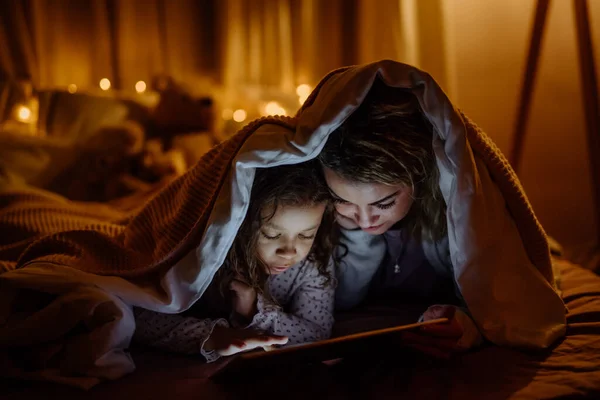 Madre feliz con su pequeña hija acostada debajo de la manta y viendo películas en la tableta en casa. — Foto de Stock