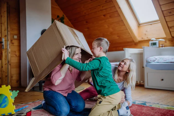 Niño con síndrome de Down con su madre y su abuela jugando con la caja juntos en casa. — Foto de Stock