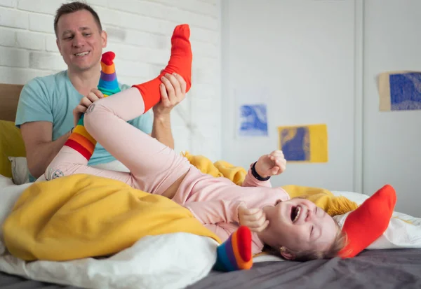 Allegra bambina con la sindrome di Down sdraiata sul letto e ridendo quando suo padre la solletica. — Foto Stock