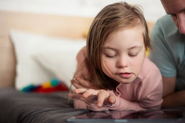 Otec a jeho malá dcera s Downovým syndromem ležící na posteli a používající doma tablet. — Stock fotografie