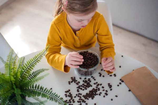 Vista de ángulo alto de niña con síndrome de Down sentada en la mesa con un tazón lleno de granos de café en casa. — Foto de Stock