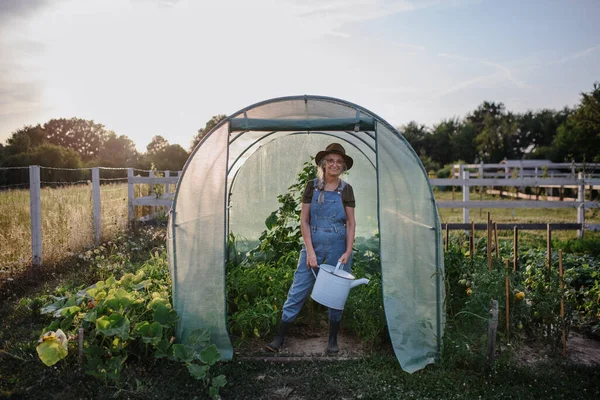 Senior tuinman vrouw met gieter in kas op de tuin, op zoek naar camera. — Stockfoto