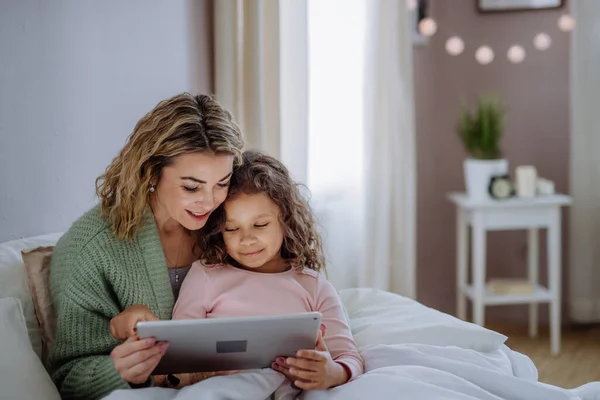 Madre feliz con su pequeña hija acostada en la cama y usando la tableta en casa. — Foto de Stock