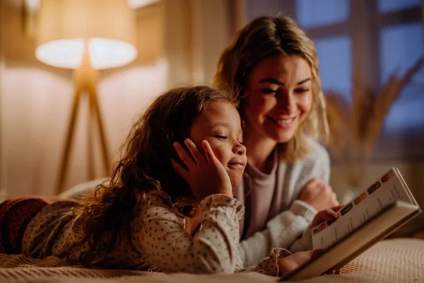 Щаслива мати зі своєю маленькою донькою лежить на ліжку і читає книгу ввечері вдома . — стокове фото