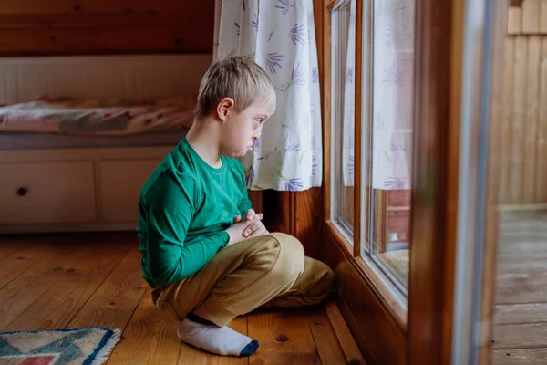 Sorglig liten pojke med Down syndrom sitter på golvet och tittar genom fönstret hemma. — Stockfoto