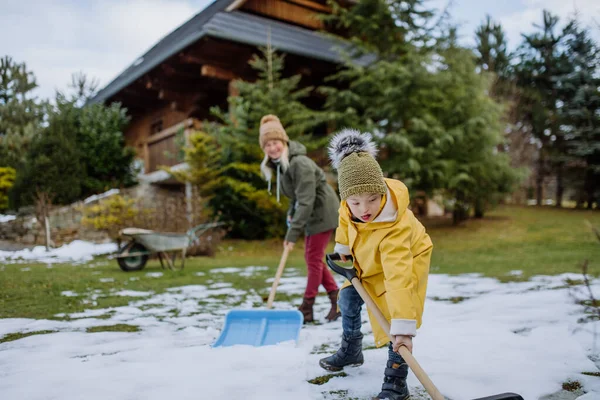 Niño con síndrome de Down con su madre limpiando la nieve del camino con pala en frente de la casa. —  Fotos de Stock