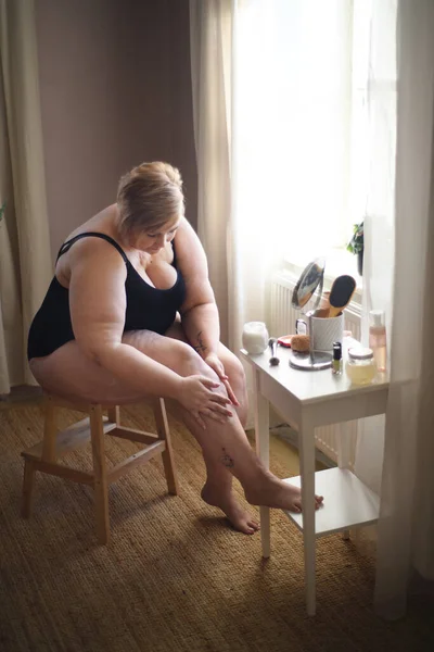 Mujer con sobrepeso sentada y aplicando crema en sus piernas en casa, concepto de autocuidado. — Foto de Stock