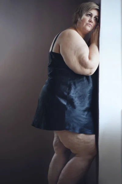 Deprimida mulher gorda solitária de pé e olhando pela janela em casa. — Fotografia de Stock