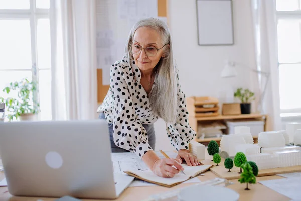 Senior vrouwelijke architect met model van huizen in kantoor en het schrijven van notities. — Stockfoto