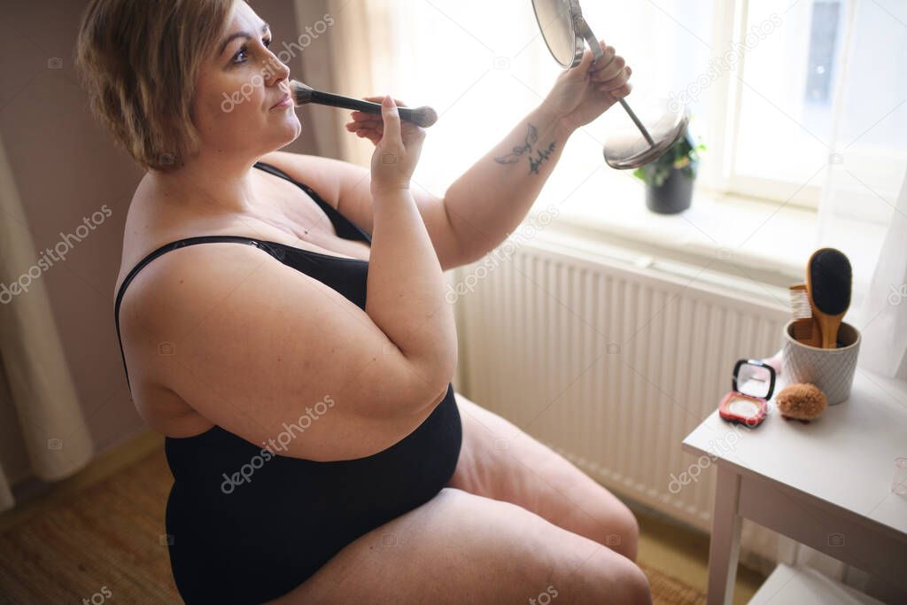 Overweight woman sitting and looking at mirror at home, selfcare concept.