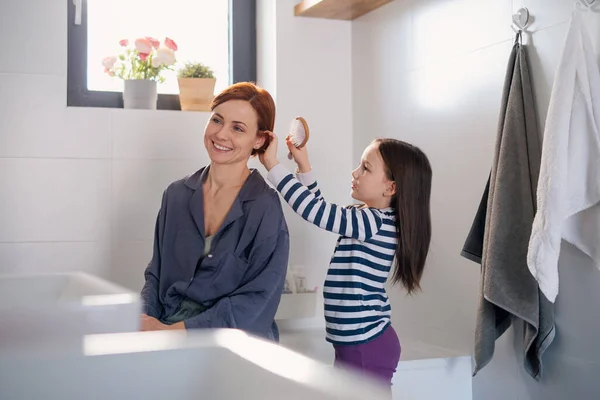 Klein meisje borstelen haar moeders haar in de badkamer, ochtend routine concept. — Stockfoto