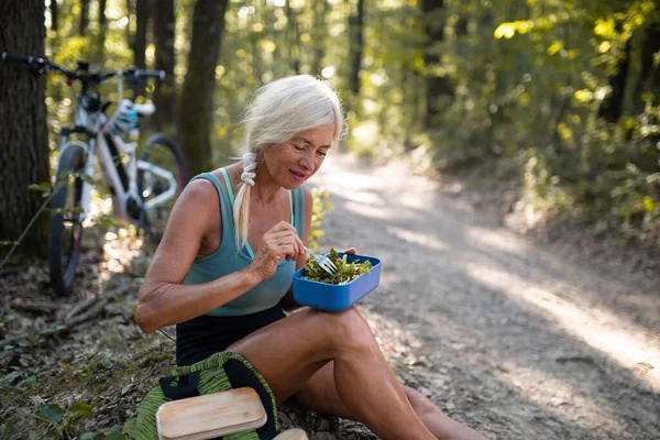 Glückliche Seniorin rastet aus, sitzt und isst Imbiss im Wald. — Stockfoto