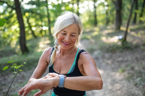 Ritratto di donna anziana attiva corridore in piedi all'aperto nella foresta, impostazione smartwatch. — Foto Stock