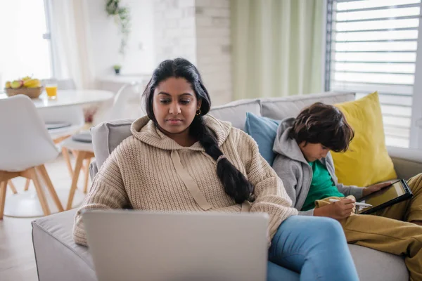 India madre usando el ordenador portátil y su hijo pequeño usando tableta y sentado en el sofá en casa. — Foto de Stock