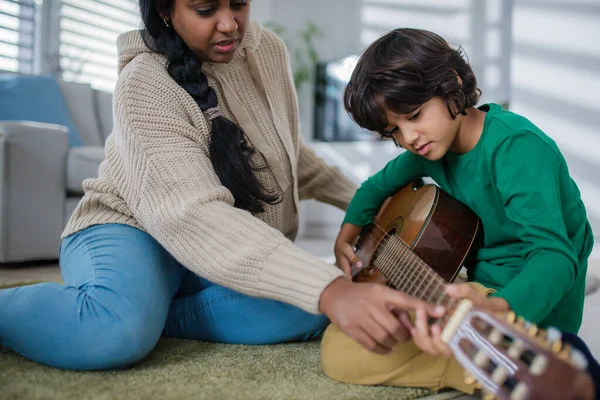 Piccolo ragazzo multirazziale che impara a suonare la chitarra con sua madre a casa. — Foto Stock
