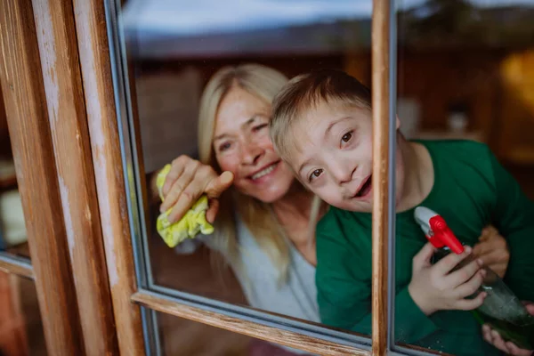 Chłopiec z zespołem Downa z babcią sprzątającą okno w domu. — Zdjęcie stockowe