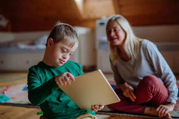 Kind mit Down-Syndrom sitzt auf dem Boden und verwendet Tablette mit Großmutter zu Hause. — Stockfoto